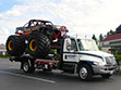 A monster truck with huge tires is being hauled on a Ron May Towing flatbed towi truck